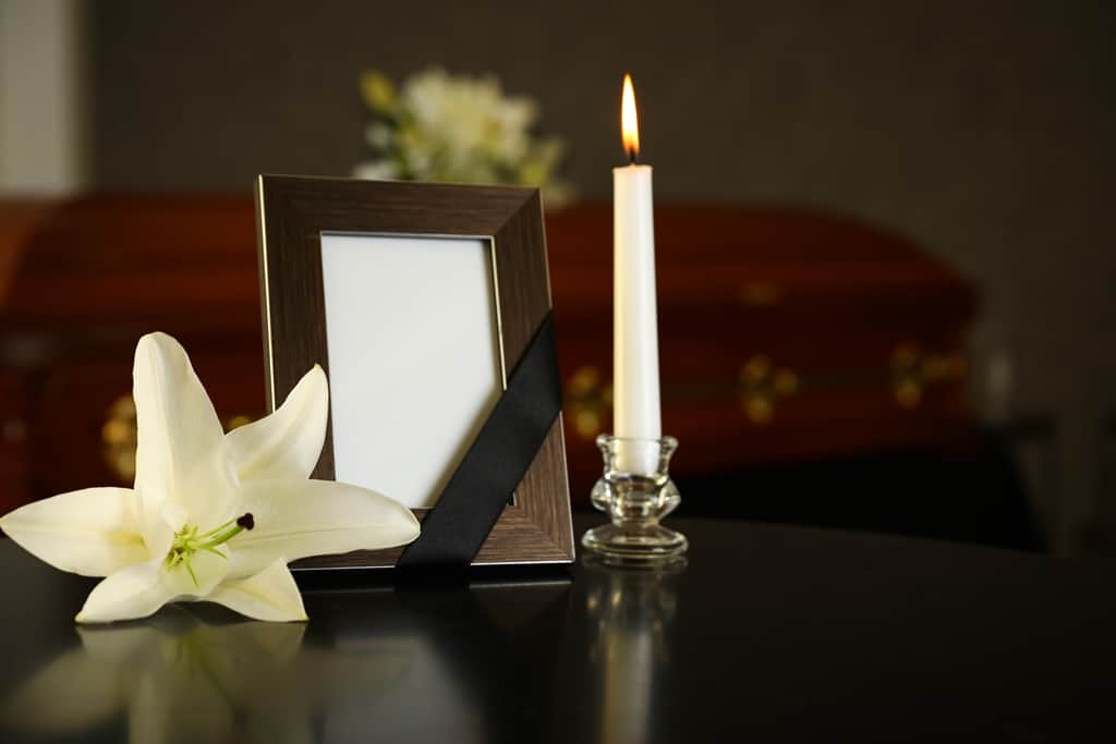 Ramka na zdjęcie z żałobną wstęgą, lilia i zapalona świeca obok trumny